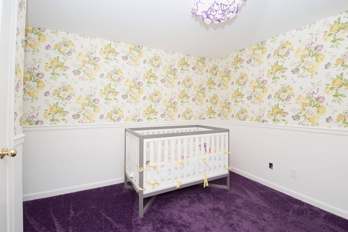 Ein Zimmer mit floralem Wandpapier, einem Babybett und violettem Teppich.