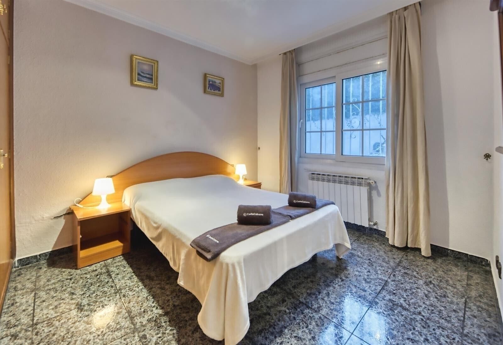 Ett hotellrum med en säng, två sänglampor, ett fönster med gardiner och ett element.