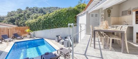 Ein Pool mit Liegestühlen, ein Essbereich mit Stühlen und Blick auf einen bewaldeten Hügel.