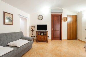 Una sala de estar con un sofá gris, un televisor sobre un mueble de madera, un reloj en la pared y una puerta de madera.