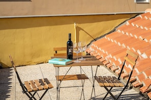 Una mesa con una botella, vasos y un libro en una terraza con sillas de madera y techo de baldosas.