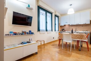 Una cocina moderna con armarios blancos, un comedor con mesa y sillas, un televisor en la pared y un taburete pequeño.