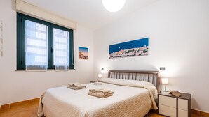 A bedroom with a bed, two bedside tables, a nightstand, a window with blinds, and a wall-mounted painting. - Vico 1° Sulis Apartment 3 with Wi-Fi and Air Conditioning (Cagliari)