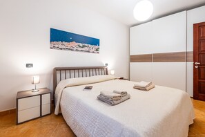 Un dormitorio con cama, lámparas de noche, mesita de noche, una toalla doblada, un cuadro de un paisaje urbano y un armario.