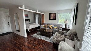 A compact living space with a bed, sofa, and coffee table, featuring a wooden floor and a window with blinds.