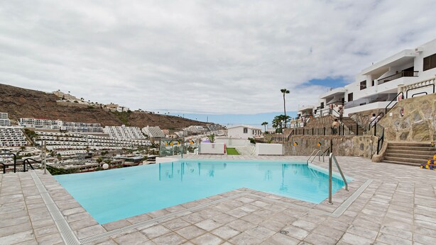 A swimming pool with a tiled deck, a stone wall, and a staircase leading to a building. - Vacation Apartment 'Aloe Puerto Rico Beach' with Sea View, Wi-Fi and Air Conditioning (Puertorico -Mogan)
