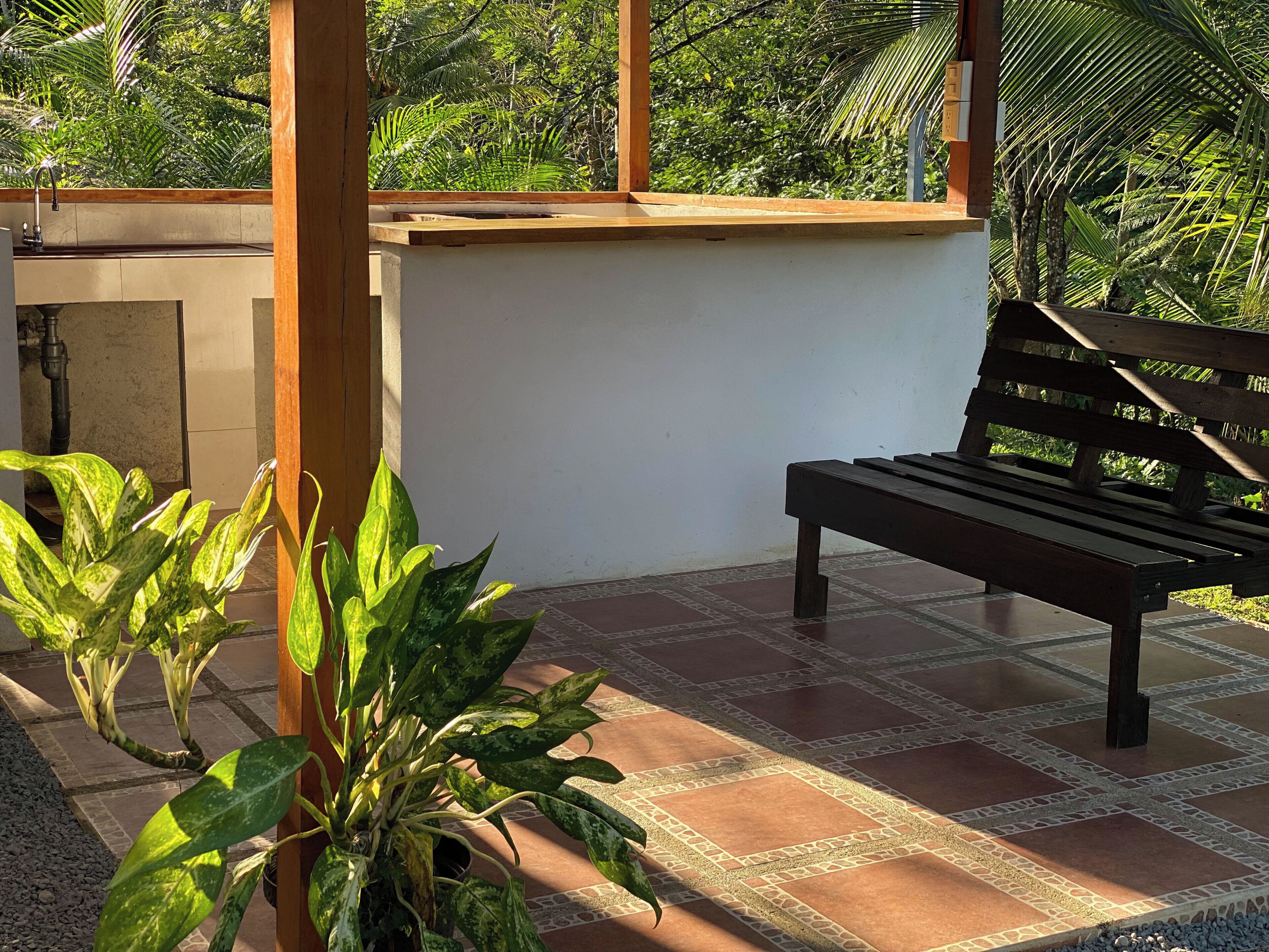 Um banco de madeira numa varanda ensolarada, com chão em mosaico e plantas em vasos.