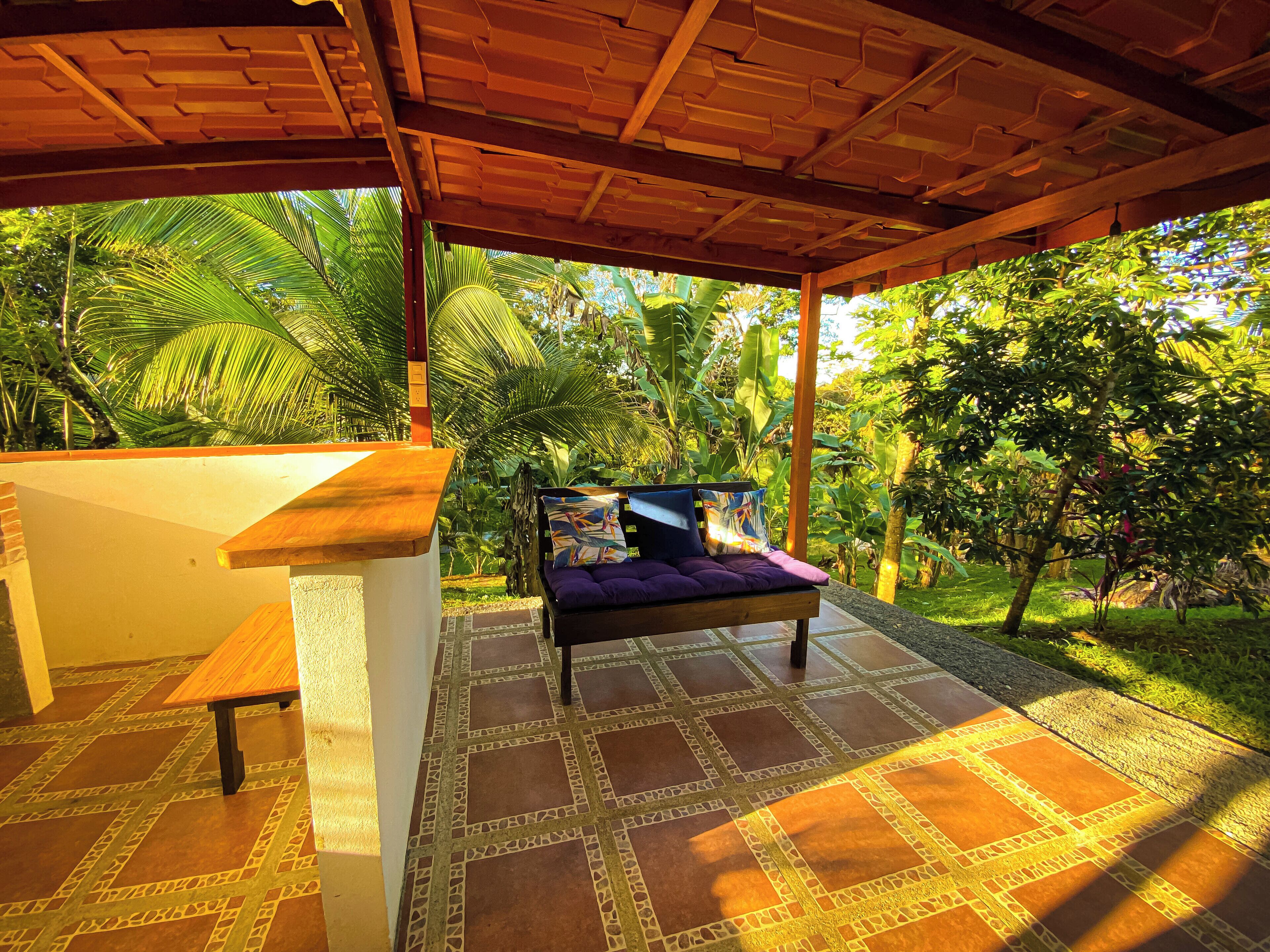 A covered outdoor seating area with a bench and a table, surrounded by lush greenery and a patterned floor.