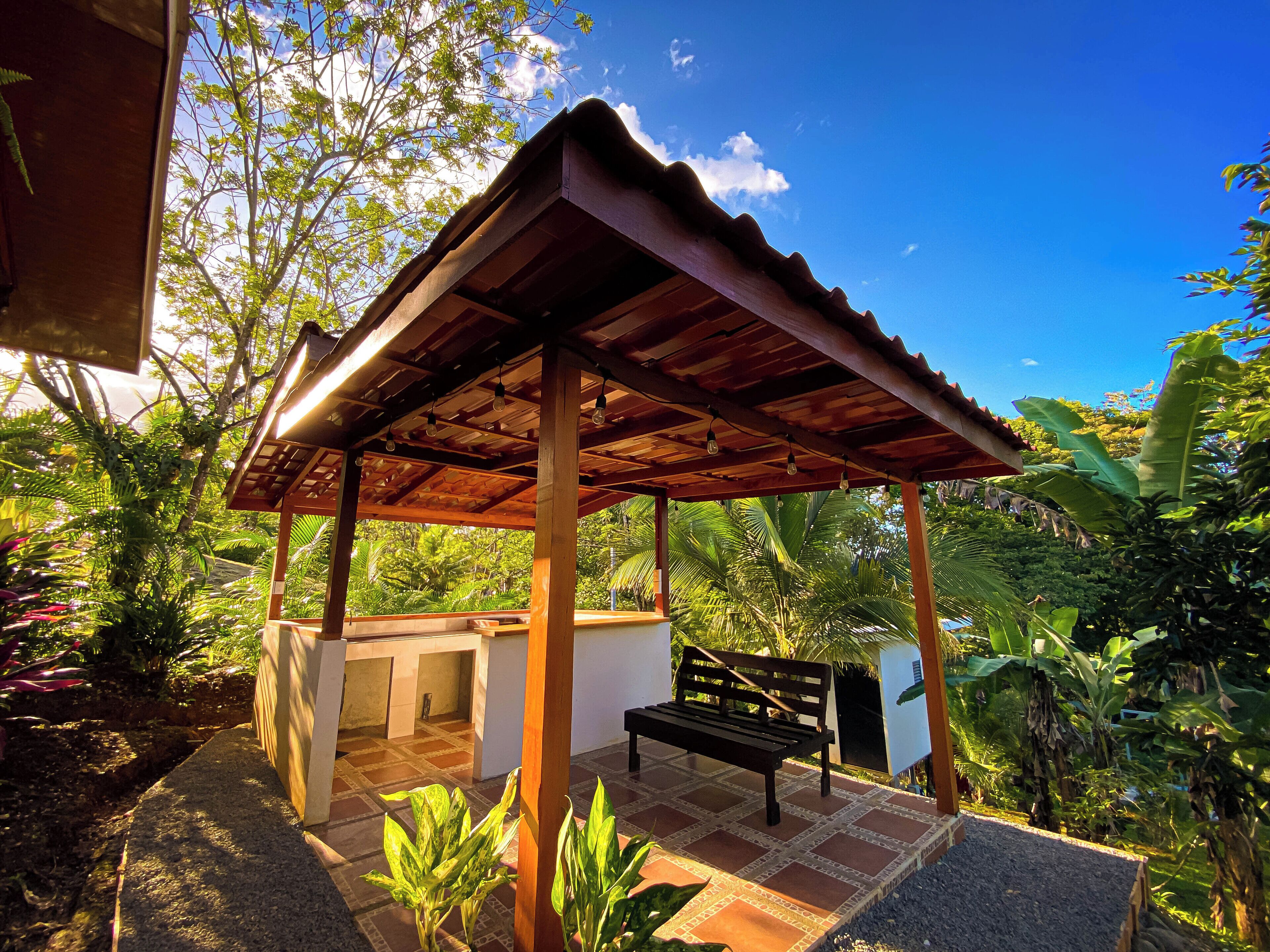 A covered outdoor seating area with a bench and a small table, surrounded by lush greenery and a clear blue sky.