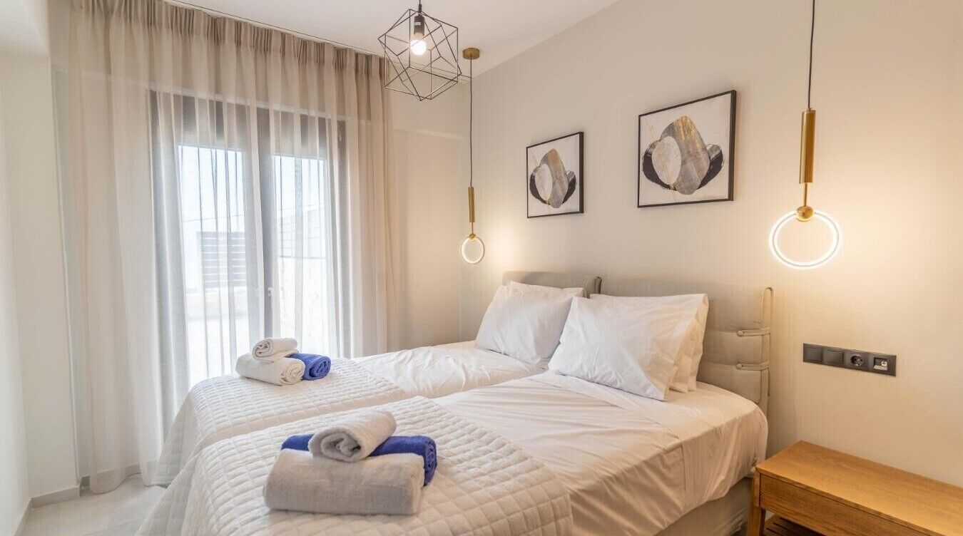 A hotel room with two beds, a bedside table, a desk, a window with curtains, and wall-mounted artwork.