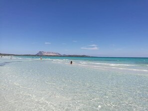 Une plage à l’eau turquoise et limpide, une montagne en arrière-plan et quelques personnes au loin.