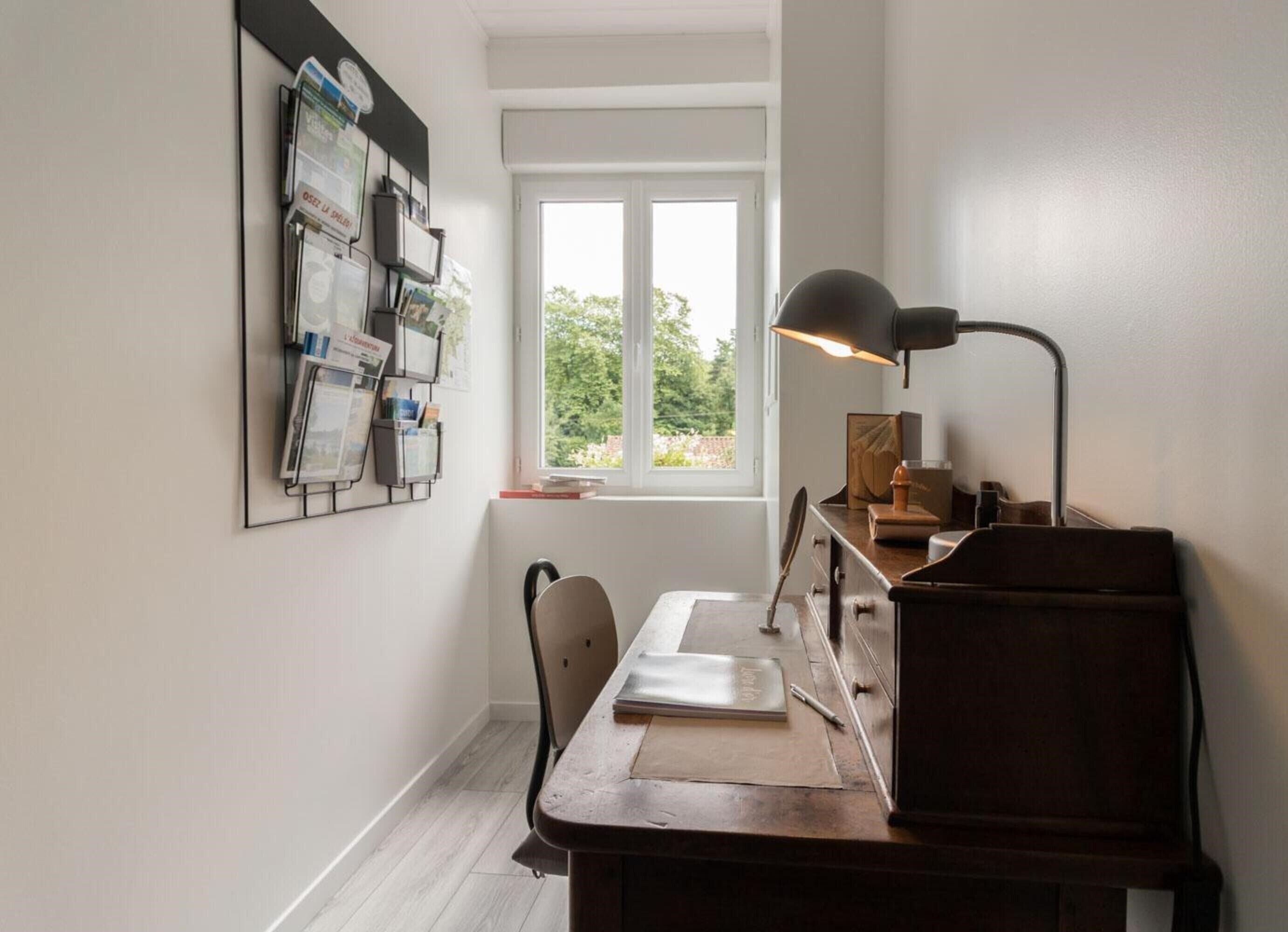 Et værelse med skrivebord, stol, lampe og et vindue med udsigt til grønne områder.