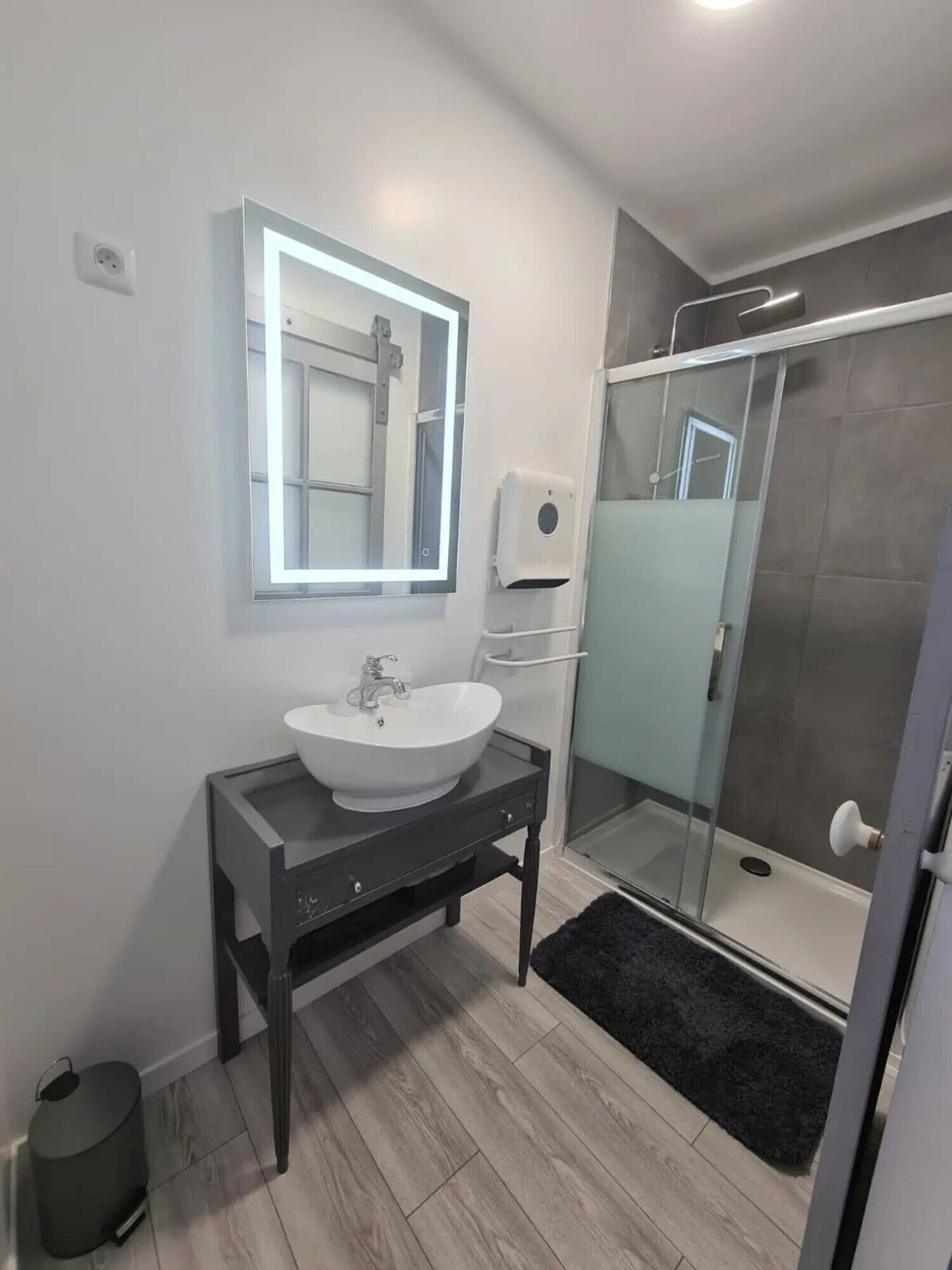 A modern bathroom with a white sink, a mirror with built-in lighting, a glass shower enclosure, and a black rug.