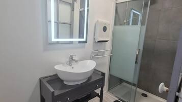 A modern bathroom with a white sink, a mirror with built-in lighting, a glass shower enclosure, and a black rug.
