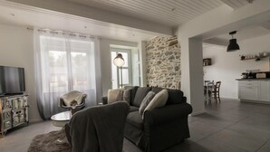 A living room with a sofa, a television, a stone wall, and a kitchen area. - Cottage 'Gîte De Pène Blanque' with Mountain View, Private Terrace and Wi-Fi (Arbas)