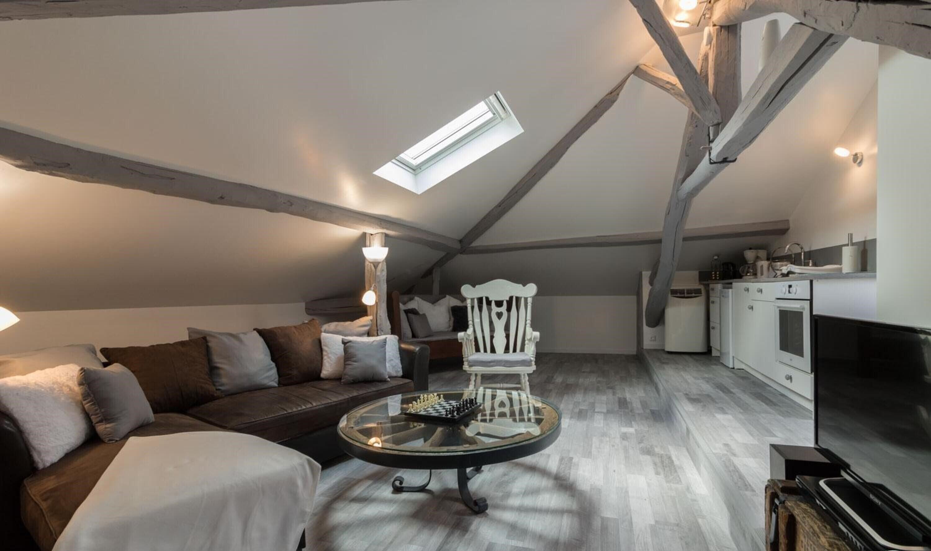 Habitación estilo loft con cama en altillo, un sofá, una mesa de centro de vidrio, una silla blanca y una zona de cocina con estufa y fregadero.