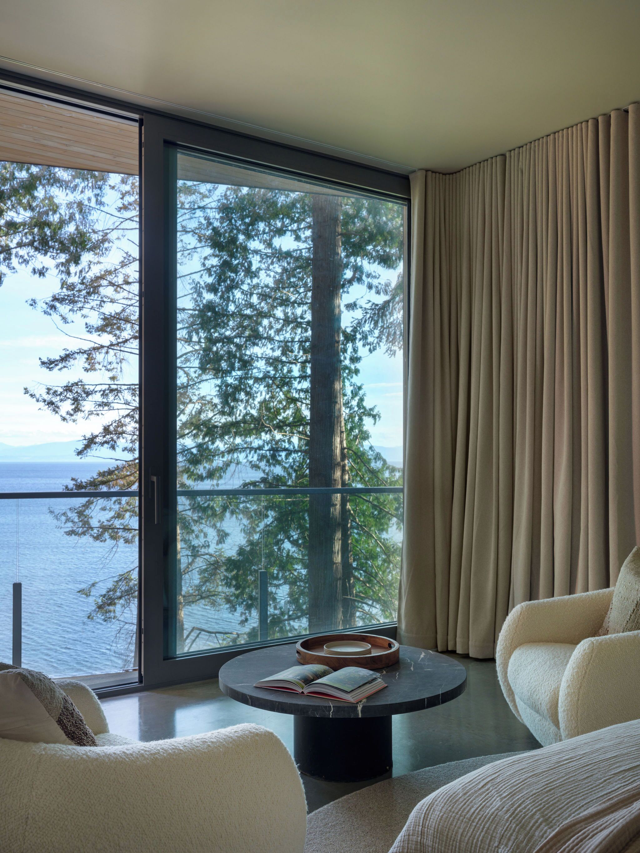Una habitación moderna con un ventanal grande que ofrece vistas a árboles y agua. Hay una mesa redonda con libros y una bandeja, y dos sillones cómodos.