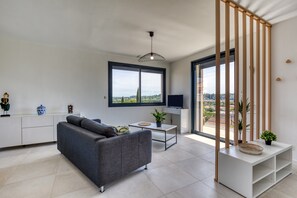 Een moderne woonkamer met een grijze bank, een klein tafeltje, een televisie en een groot raam met uitzicht op de stad.