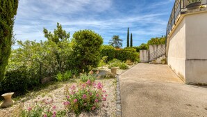 Un jardí amb un camí de grava, diferents plantes i un edifici amb mur de formigó i barana metàl·lica.