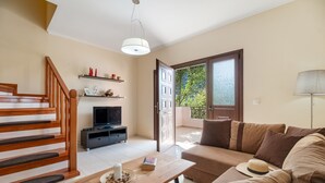 Un salon avec un escalier en bois, un canapé, une table sur laquelle se trouvent un livre et une bougie, un téléviseur et une porte donnant sur l’extérieur.