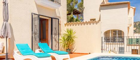 A poolside area with lounge chairs, a potted plant, and a building with a tiled roof and balcony.