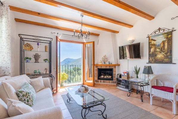 Una sala de estar con sofá, mesa de centro de vidrio, chimenea, televisión y balcón con vista a las montañas.
