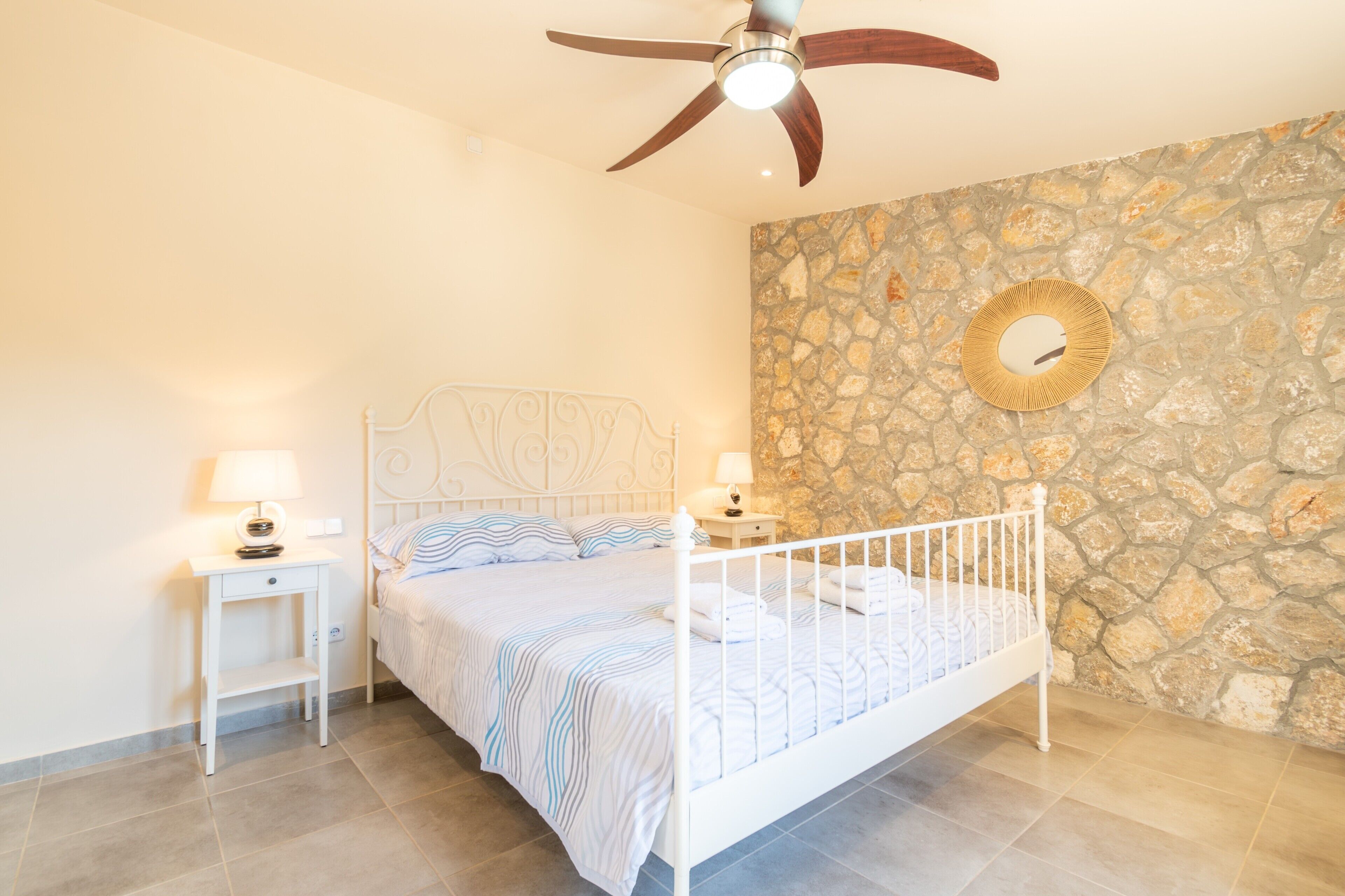 A bedroom with a white metal bed, two bedside tables with lamps, a stone wall, and a ceiling fan.