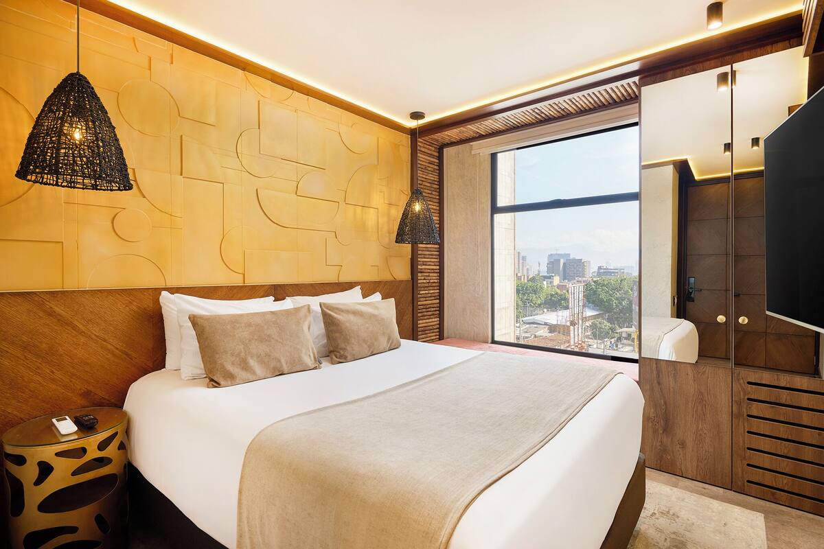 A modern hotel room with a large bed, a city view, a wooden headboard, and a TV.