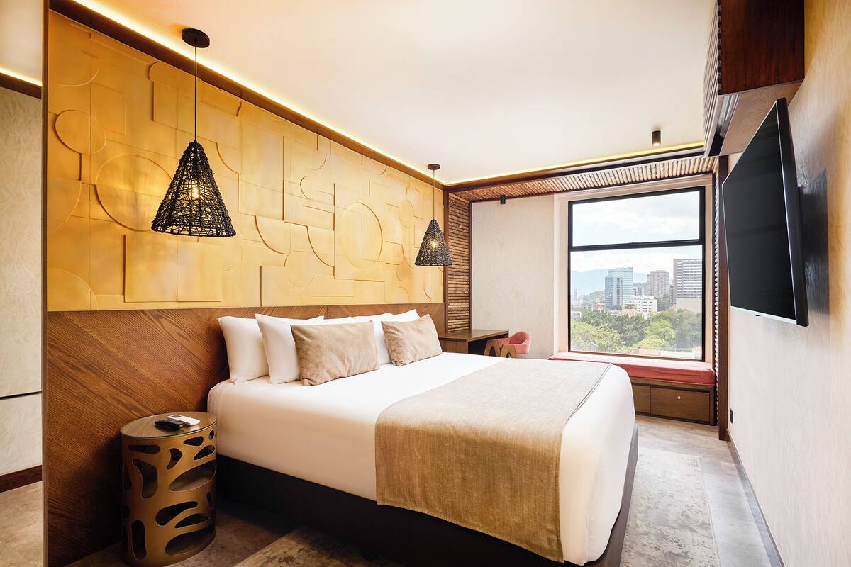 A modern hotel room with a large bed, a TV mounted on the wall, and a window with a city view.