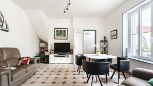 A living room with a sofa, a dining table with chairs, a television, and a door leading to another room.