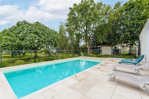 A rectangular swimming pool with a tiled deck, surrounded by a fence and trees.
