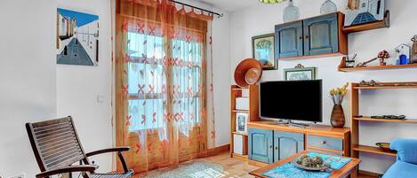 A living room with a television, a blue sofa, a wooden chair, a table with a decorative plate, and a window with a floral curtain.
