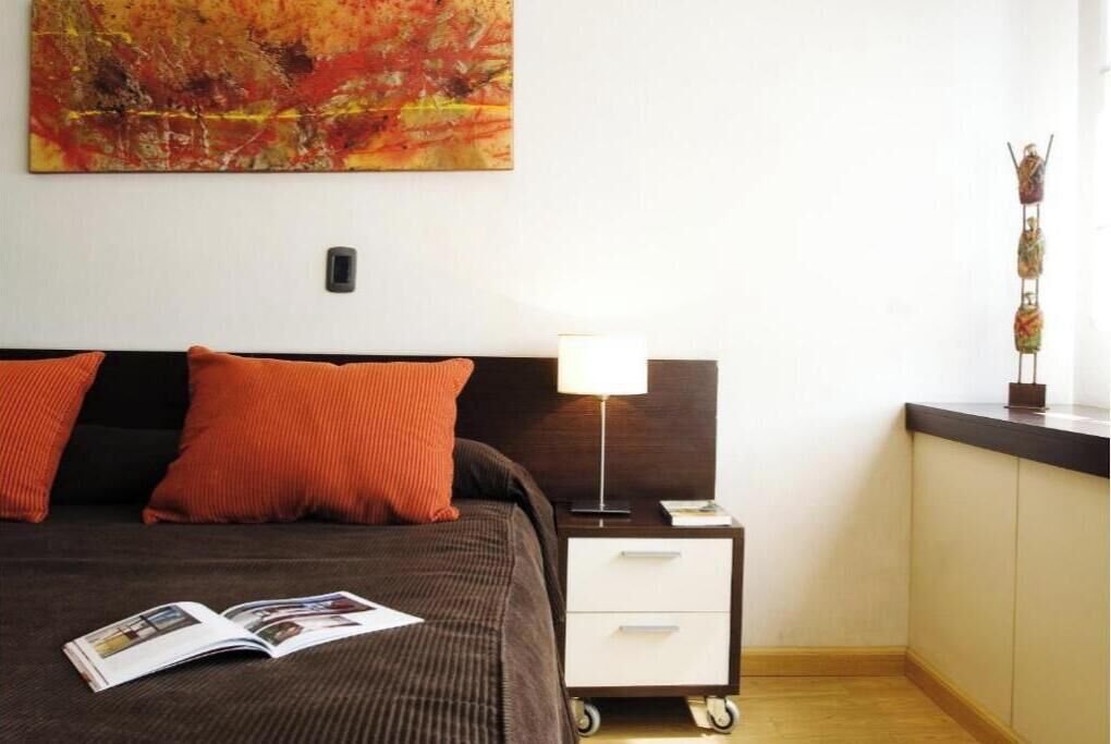 A bedroom with a bed, a nightstand, a lamp, a magazine, and a wall painting.