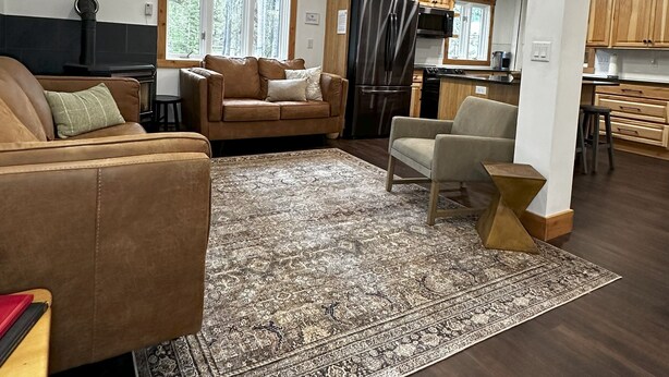 A spacious living area with a large rug, a sofa, a coffee table, and a dining area with wooden cabinets and a refrigerator. - Family Retreat 29 Miles to Yellowstone in a Quiet Wooded Area, with a Kids Nook (Island Park)
