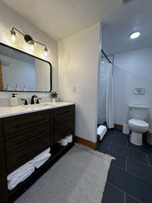 Ein modernes Badezimmer mit einem dunklen Holz-Waschtisch, einem großen Spiegel und einem weißen Waschbecken. Ein Duschvorhang und eine Toilette sind im Hintergrund zu sehen.