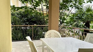 Un balcón con una mesa y sillas blancas, rodeado de follaje y una barandilla de metal.