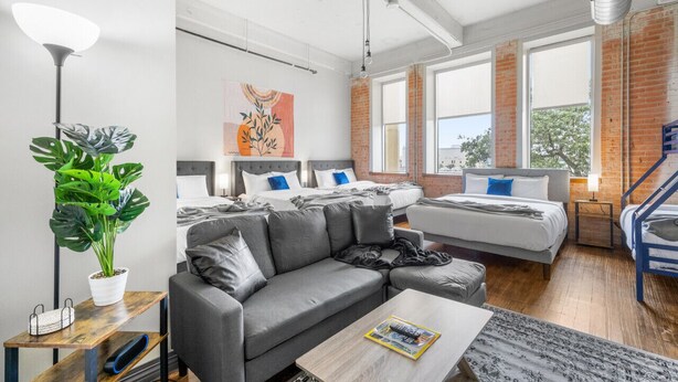 A modern hotel room with a grey sofa, two beds, a large window, and a staircase. - Spacious Downtown Apartment | Pool + In-Unit laundry (Dallas)
