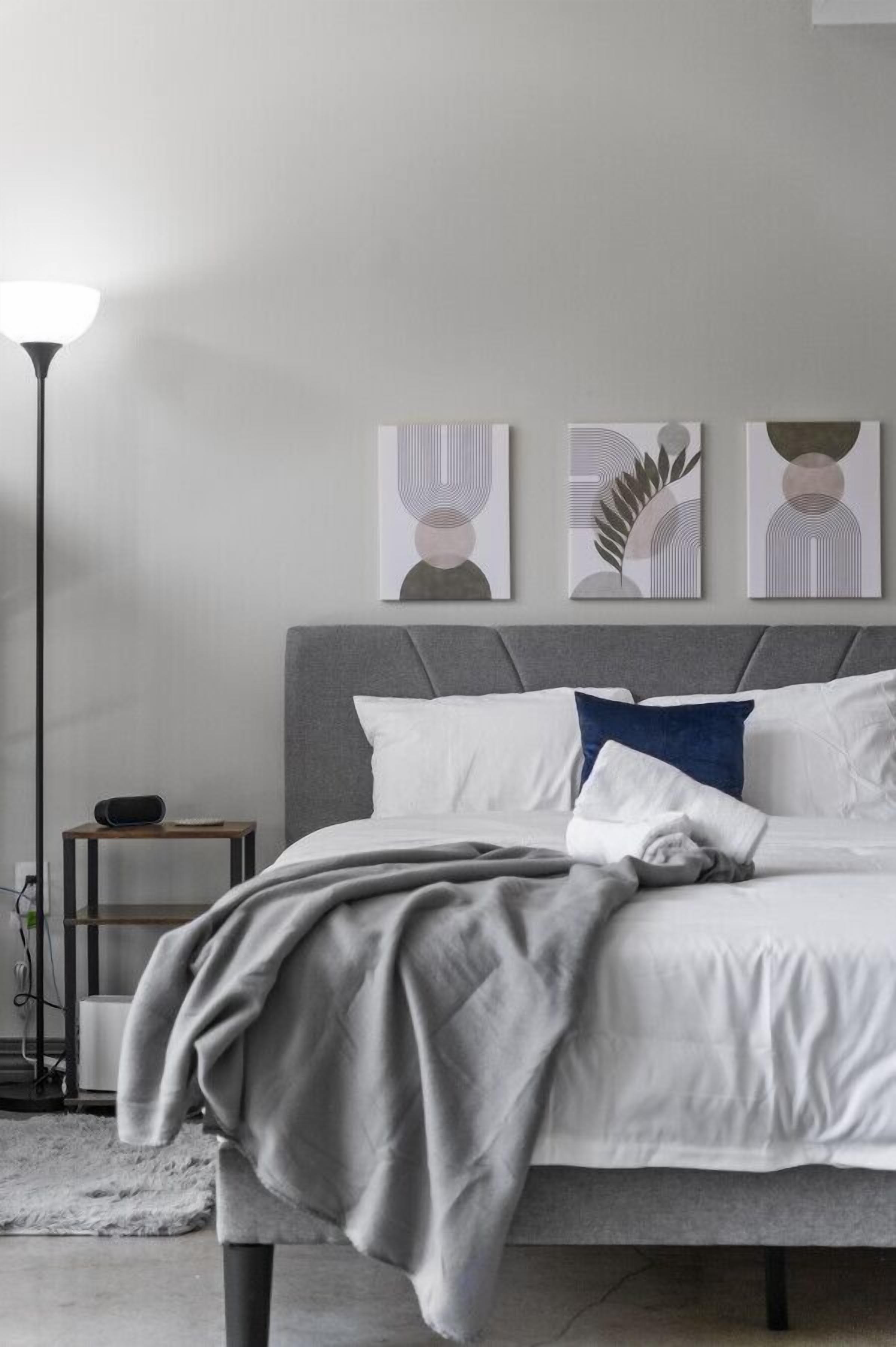 Un dormitorio moderno con una cama tapizada en gris, almohadas blancas y azules, un plaid gris y tres piezas de arte abstracto en la pared.