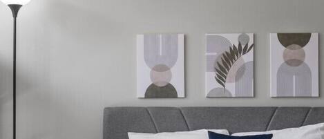 Un dormitorio moderno con una cama tapizada en gris, almohadas blancas y azules, un plaid gris y tres piezas de arte abstracto en la pared.