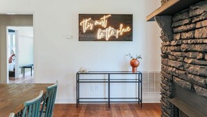 Een eetgedeelte met een houten tafel en stoelen, een neonbord aan de muur en een stenen open haard.