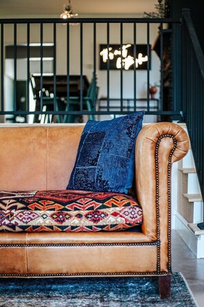 Un sofá de cuero marrón con un cojín estampado y una almohada azul, ubicado en una habitación con una escalera y un letrero de neón al fondo.