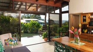 Un patio amplio con una mesa y sillas de madera, un vidrio que da a un jardín exuberante, y una zona de cocina con armarios de madera y un microondas.