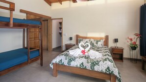 Una habitación con literas, una cama con colcha estampada, una toalla dispuesta en forma de corazón, mesitas de noche y una planta en maceta.