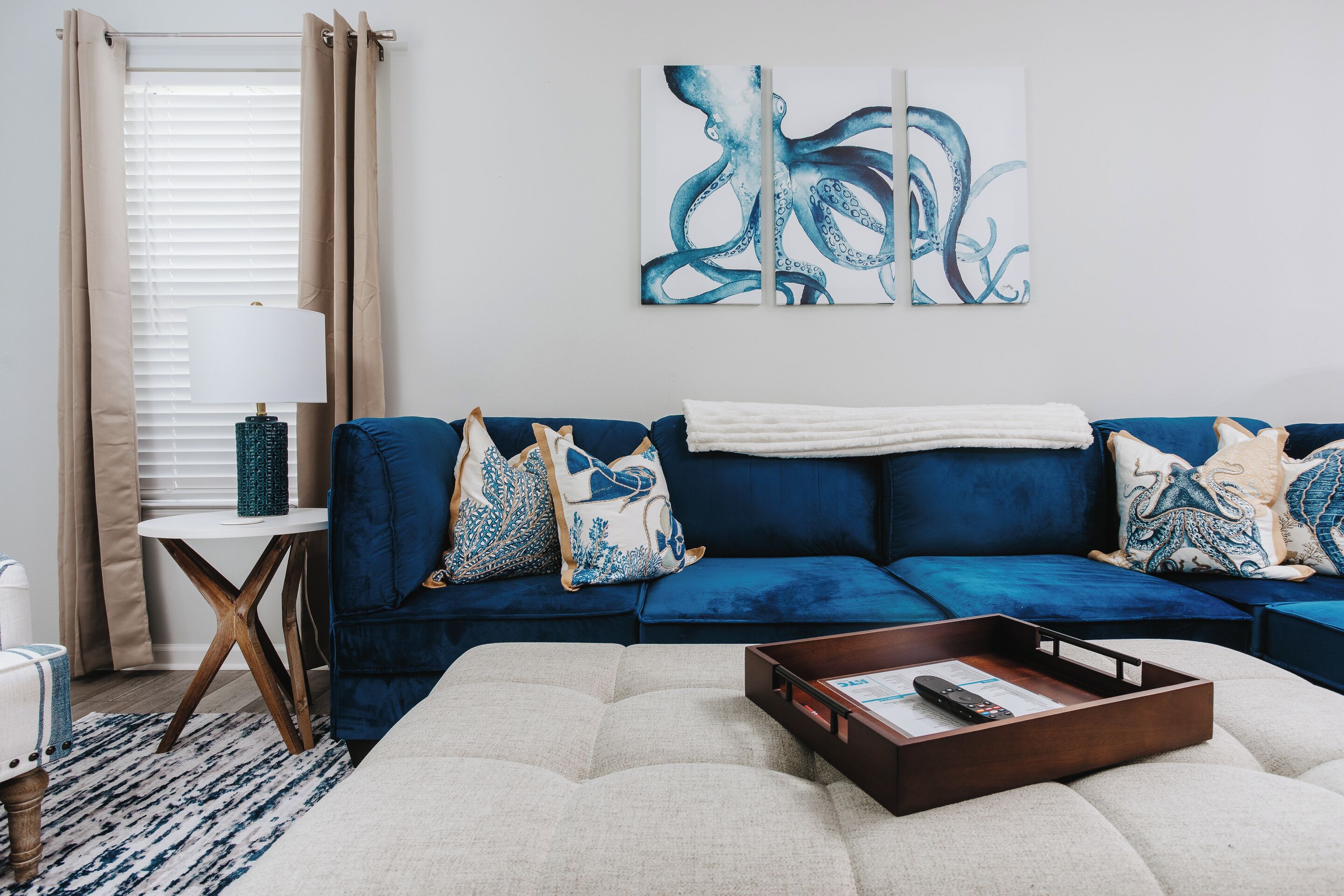 A modern living room with a blue sofa, decorative pillows, a white pouf, and a wooden side table with a lamp.