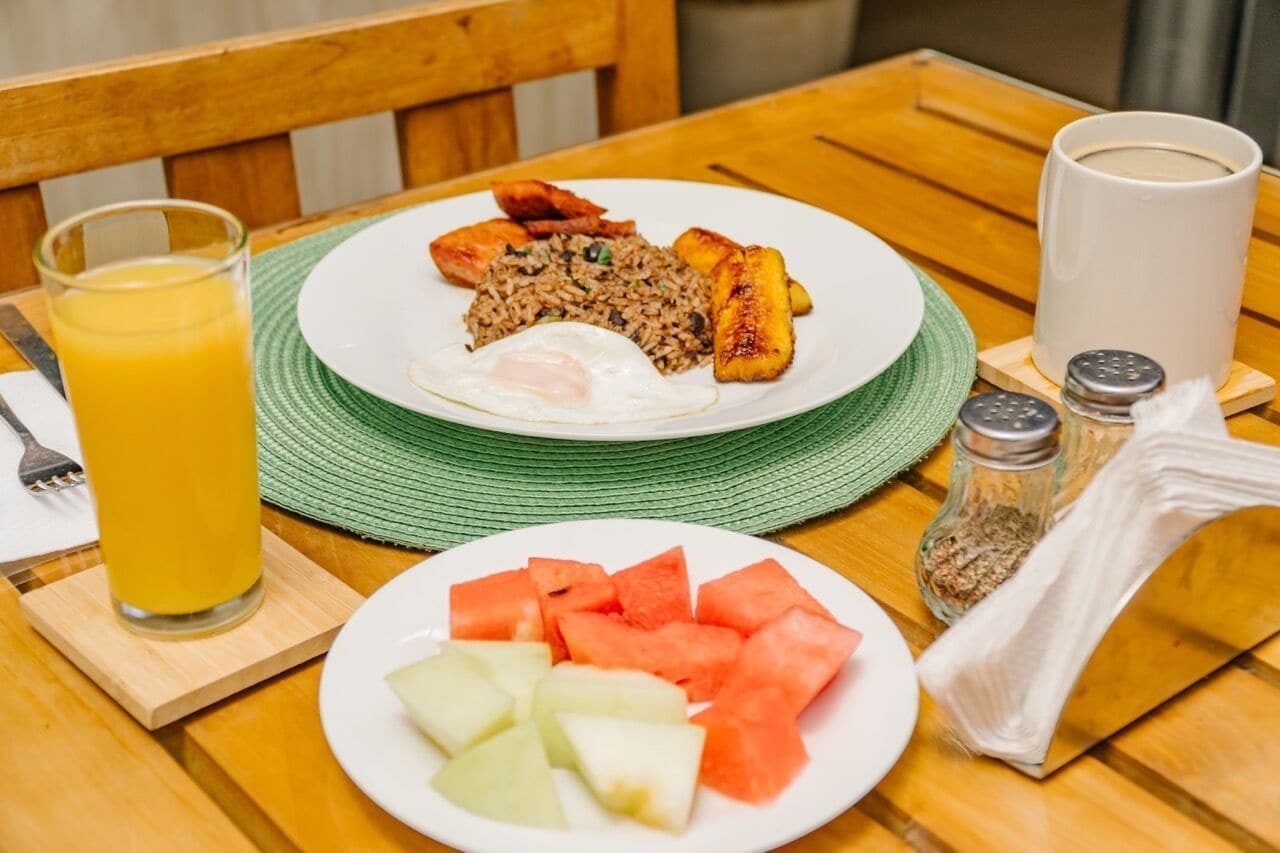 Un plato de desayuno con arroz frito, un huevo frito y plátanos maduros, acompañado de un vaso de jugo de naranja y una bandeja de sandía y melón.