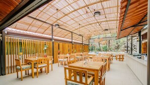 Un amplio comedor al aire libre con mesas y sillas de madera, techo de bambú y vista a zonas verdes.