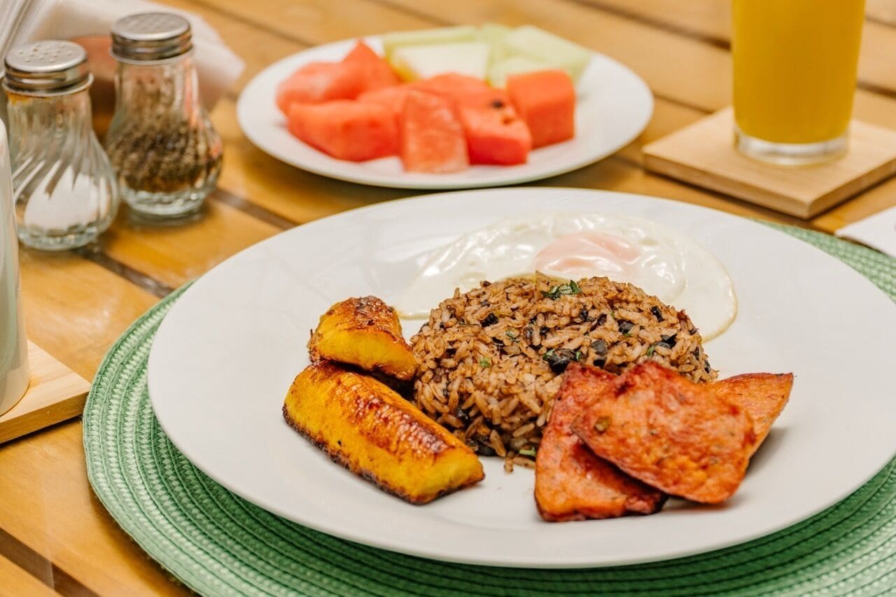 A plate of fried rice with vegetables and two fried plantains, accompanied by a slice of meat and a glass of orange juice.