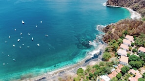 An aerial view of a coastal area with a clear blue sea, several boats, and a row of houses with red roofs along the shore. - Hotel Ocotal B&B (Sardinal)