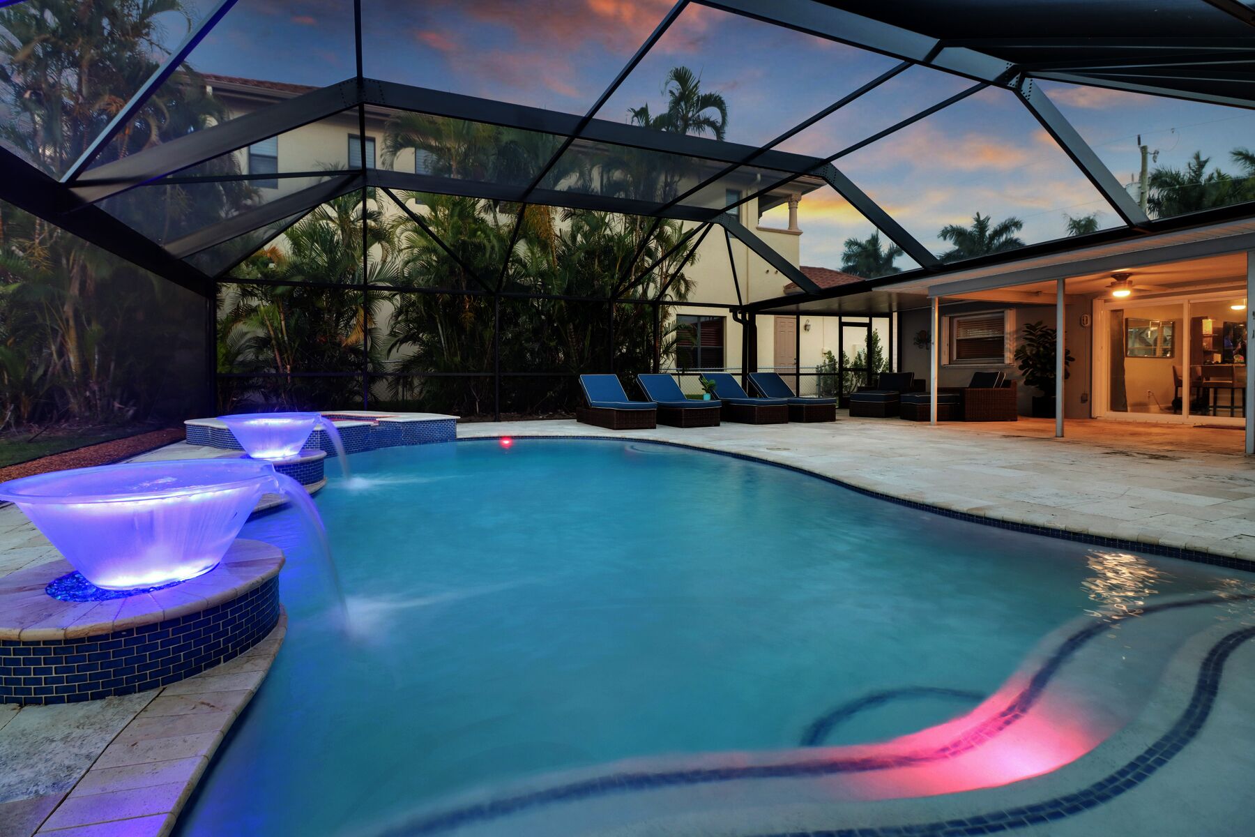 A swimming pool with a glass-enclosed deck, lounge chairs, and a hot tub with blue lighting.