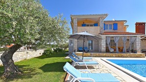 A poolside area with lounge chairs and an umbrella, a stone wall, and a building with a tiled roof. - House 2326/110968 (Istria - Vodnjan), 5000m from the beach (Vodnjan)